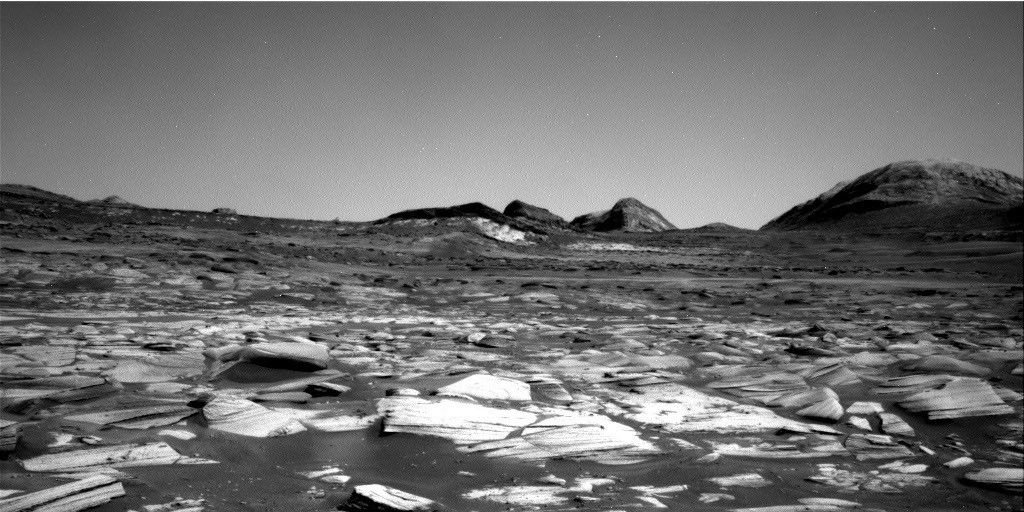 A grayscale image of the Martian surface taken by the Curiosity rover. The immediate foreground features a dense field of jagged, light-colored, thinly-layered rock fragments resting on a bed of darker sand or dust. The barren terrain gently slopes upward toward the horizon, where a cluster of dark, rugged hills and a larger, rounded mountain peak on the far right stand out against a smooth, featureless sky.