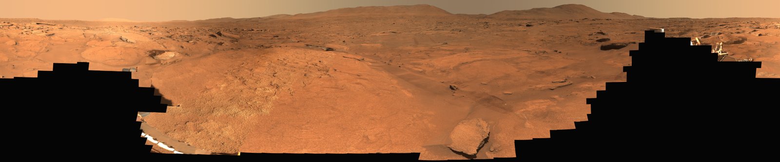A panoramic view of a barren, reddish-orange Martian landscape stretching toward distant hills, partially obscured by the jagged black silhouettes of a rover in the foreground.