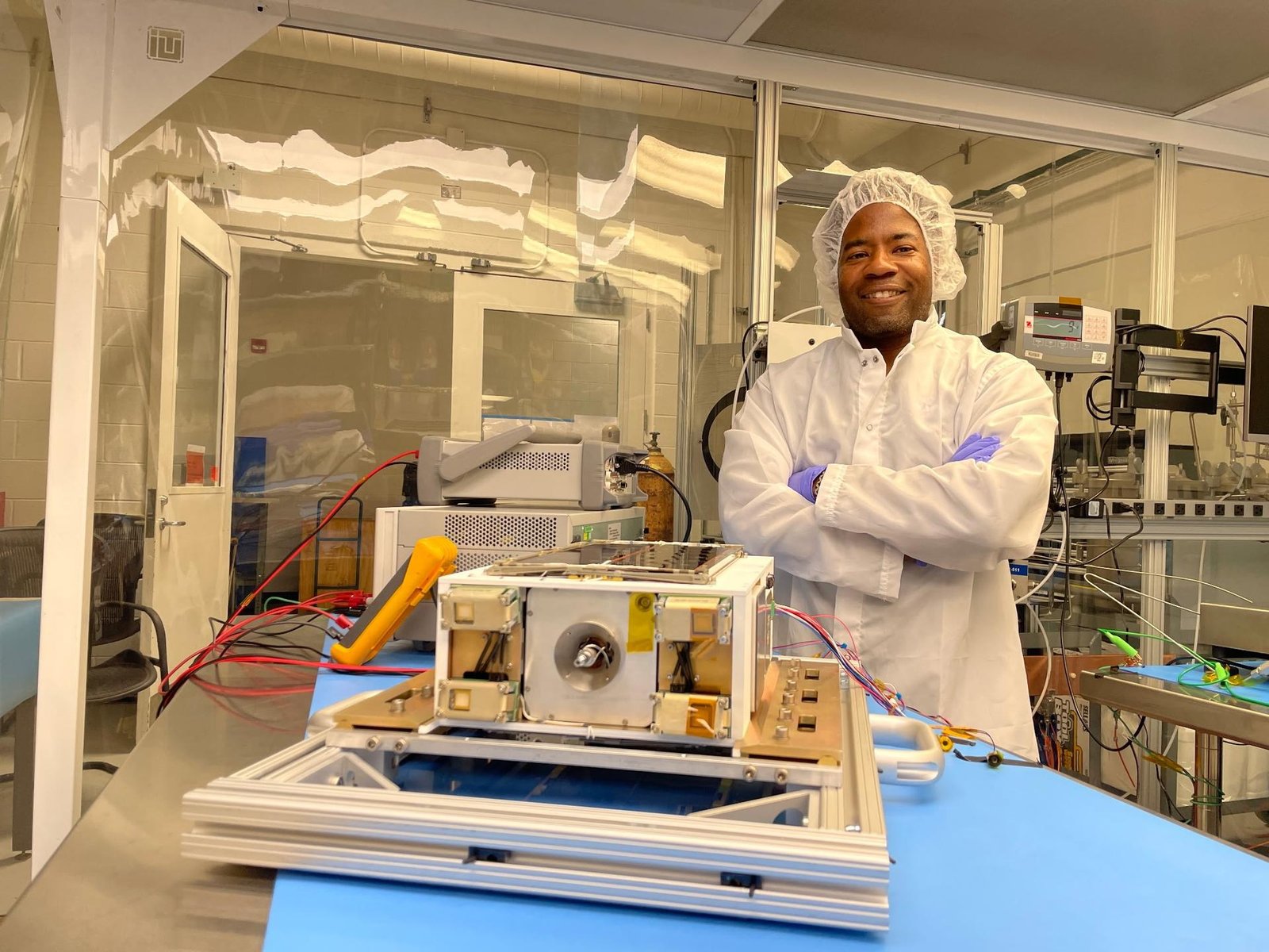 An Album of photos showing NASA engineers and technicians in white protective clean room suits working on a small, rectangular spacecraft module. They are shown inspecting the hardware on a laboratory workbench, wrapping it in silver foil, and loading it into a large, metallic vacuum chamber at NASA's Marshall Space Flight Center