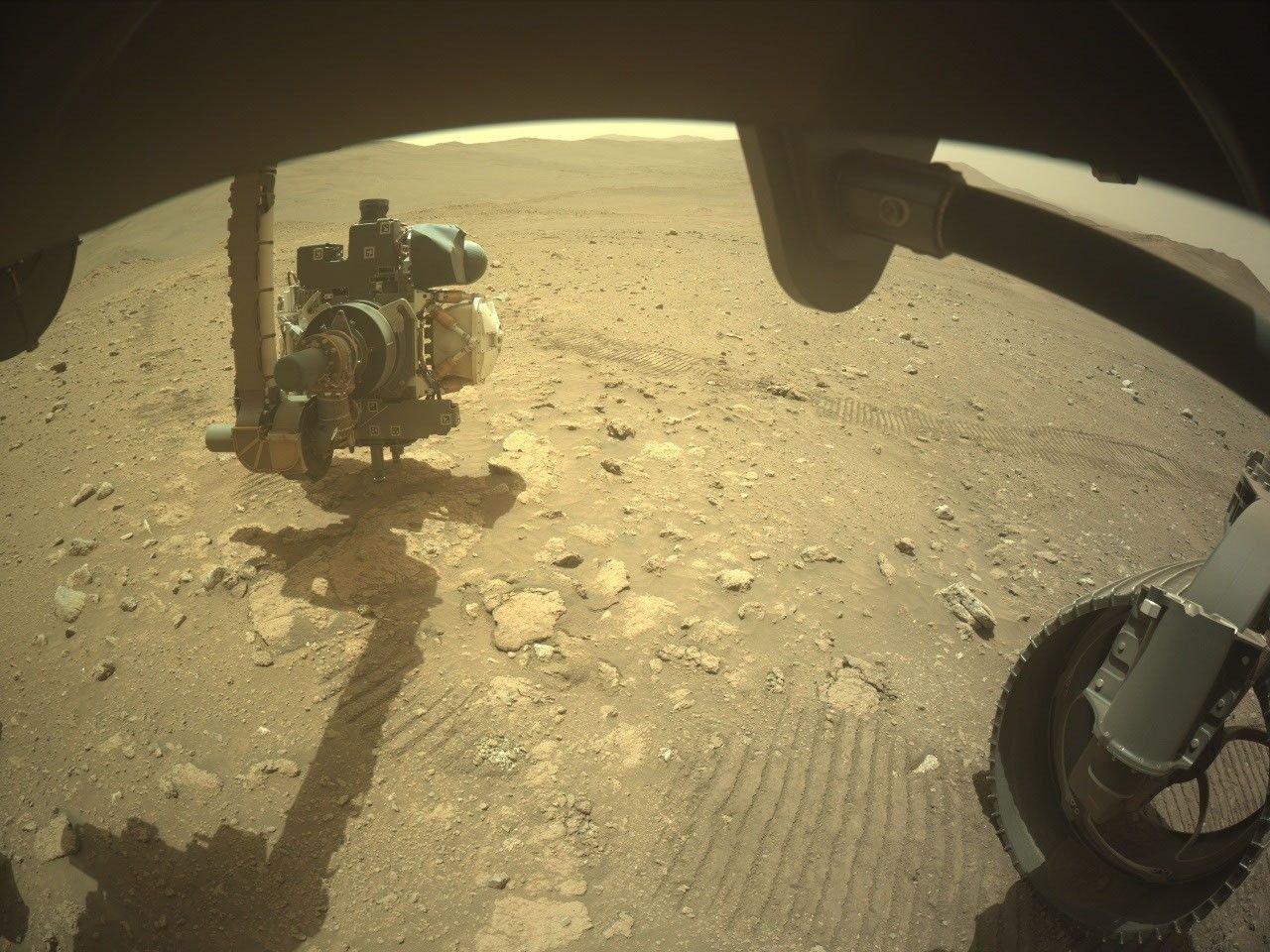 A wide-angle, slightly distorted view from beneath the Perseverance rover on Mars. Parts of the rover's dark chassis frame the top edge. On the bottom right, a section of one of its treaded wheels rests on the dirt. On the left, the rover's robotic arm, featuring a complex instrument turret at its end, is extended toward the ground. The arm casts a long, distinct shadow over the reddish-brown, rocky sand. Parallel rover tracks are pressed into the soil, leading away toward a relatively flat, barren landscape that ends at a horizon of distant, low hills under a pale sky.