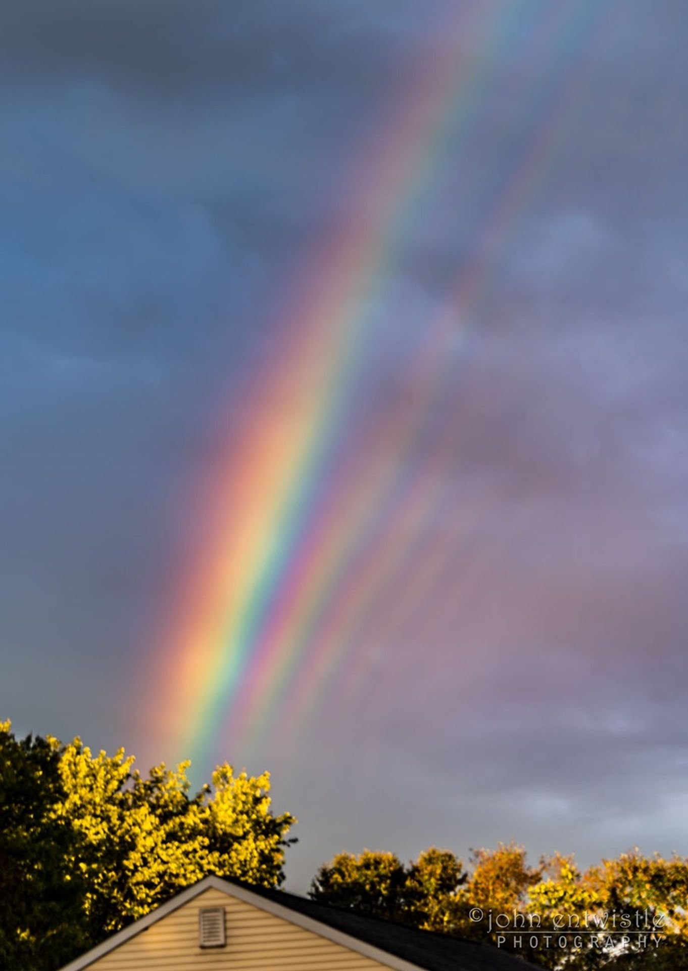 The top of a house is shown beneath a dark and cloudy sky. Many rainbow stripes are seen above the house.