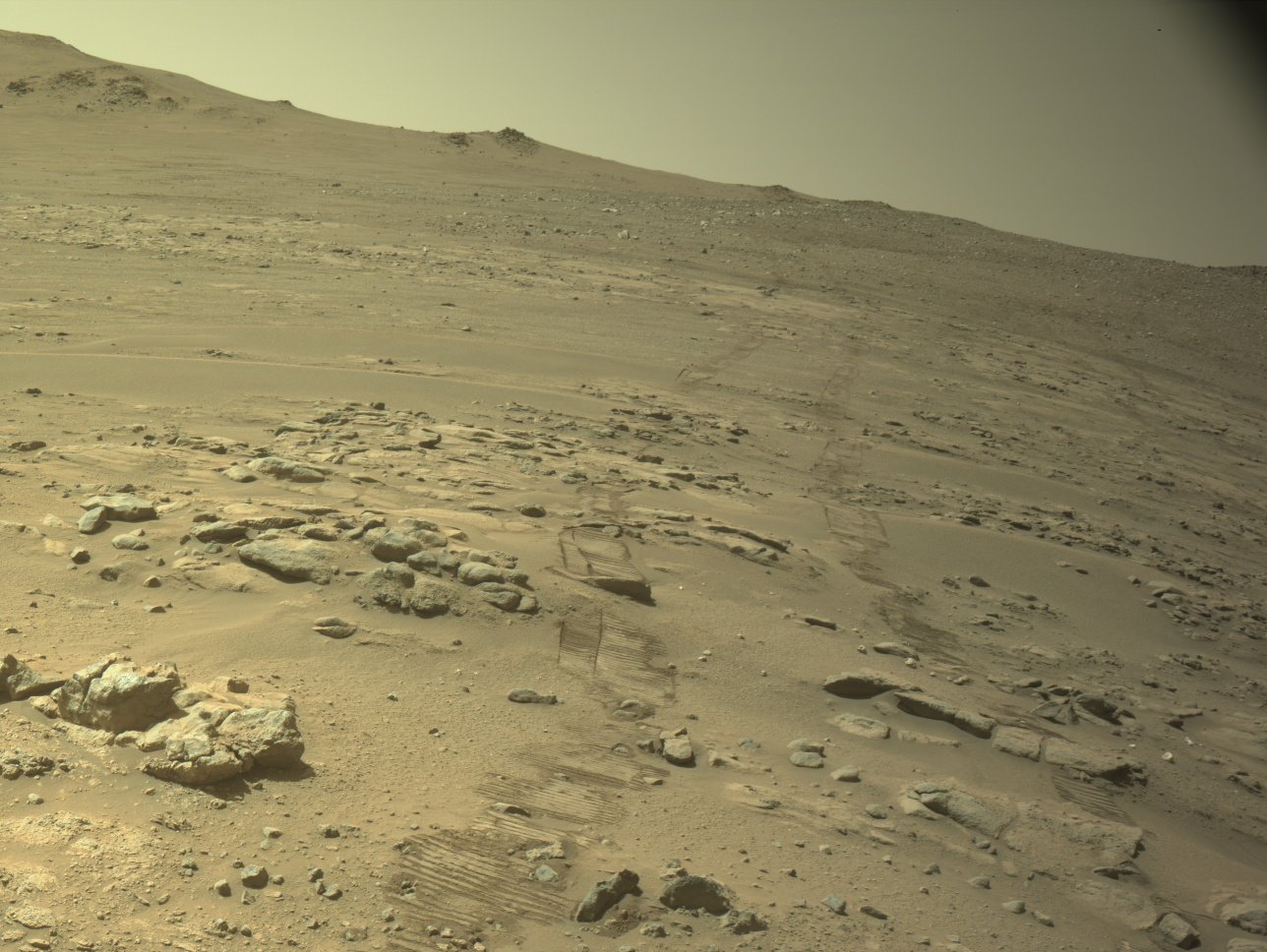 Rover tracks press into the sand of a rocky, sloping Martian landscape, leading upward toward a distant ridge under a pale, hazy sky.